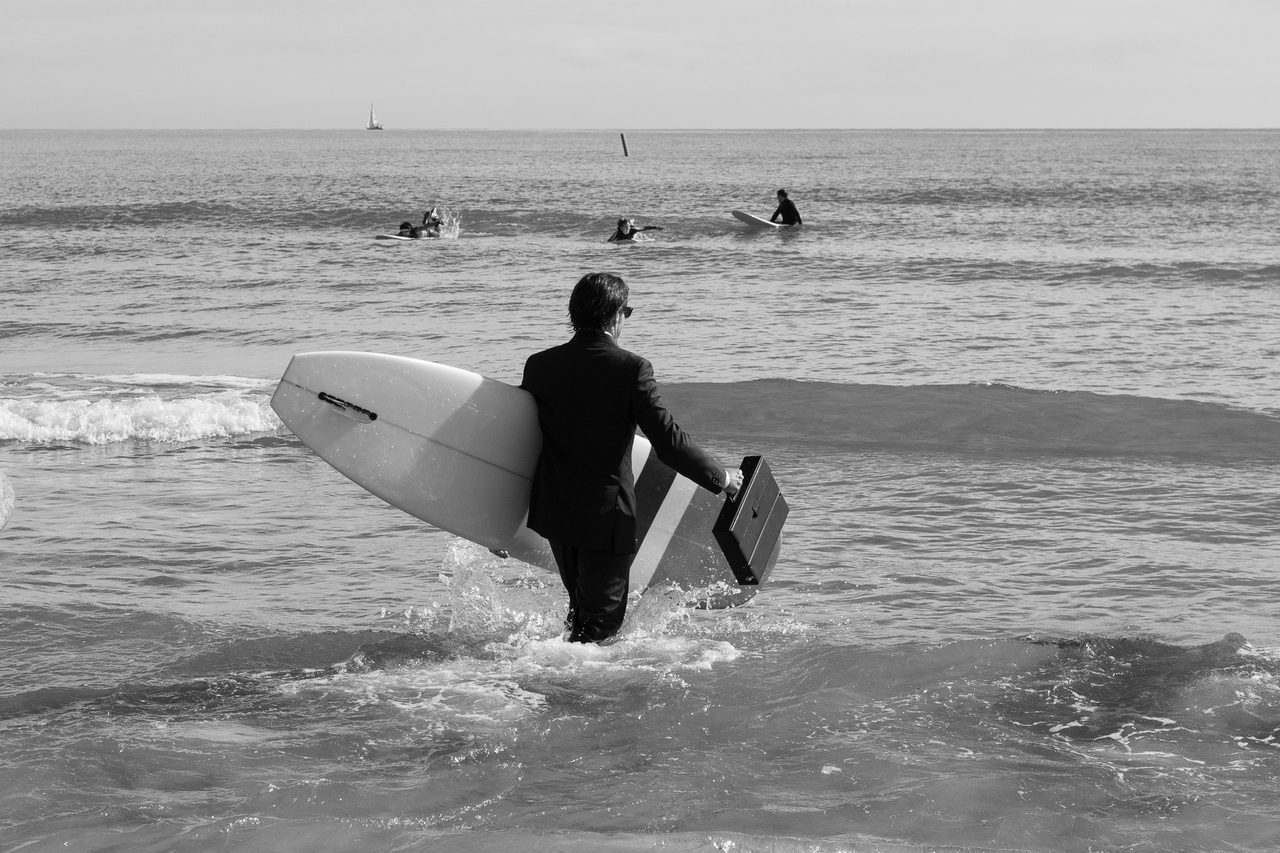 Persona vestida de ejecutivo con tabla de surf y maletín caminando hacia el mar durante el Festival Quasimoto en Burriana, Castellón, en blanco y negro.