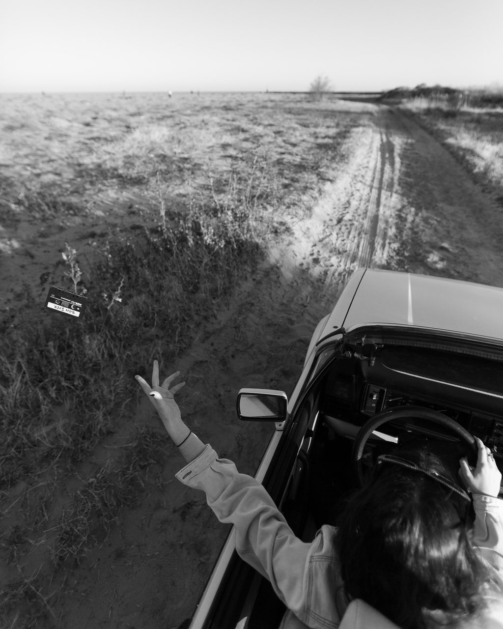 VW descapotable en un camino de arena junto a la playa de Burriana, con el conductor dejando caer una cinta de casete por la ventanilla, en blanco y negro.
