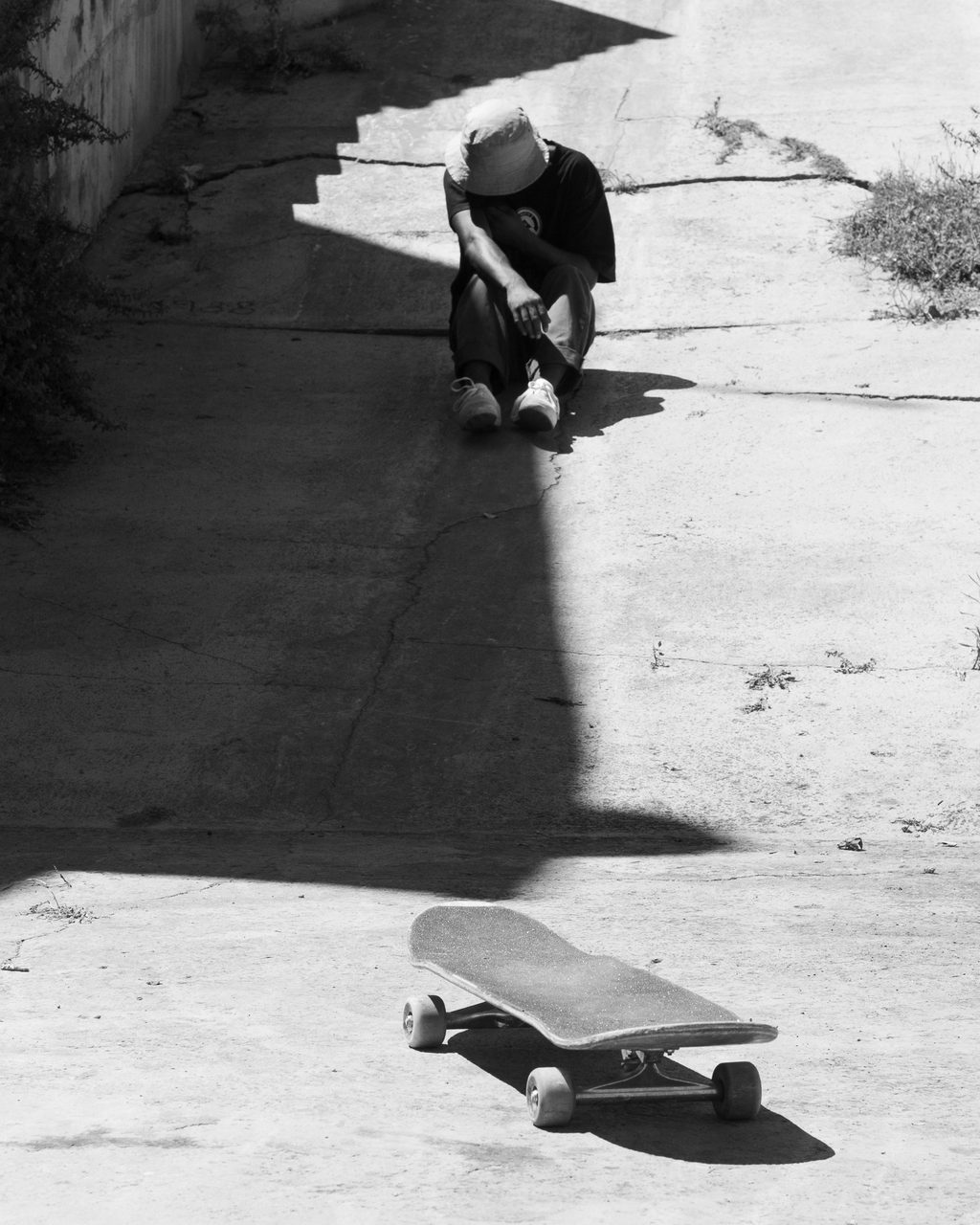 Skater sentado en la sombra de una rampa de hormigón en Castellón, con el monopatín iluminado por el sol más abajo en la pendiente, en blanco y negro.