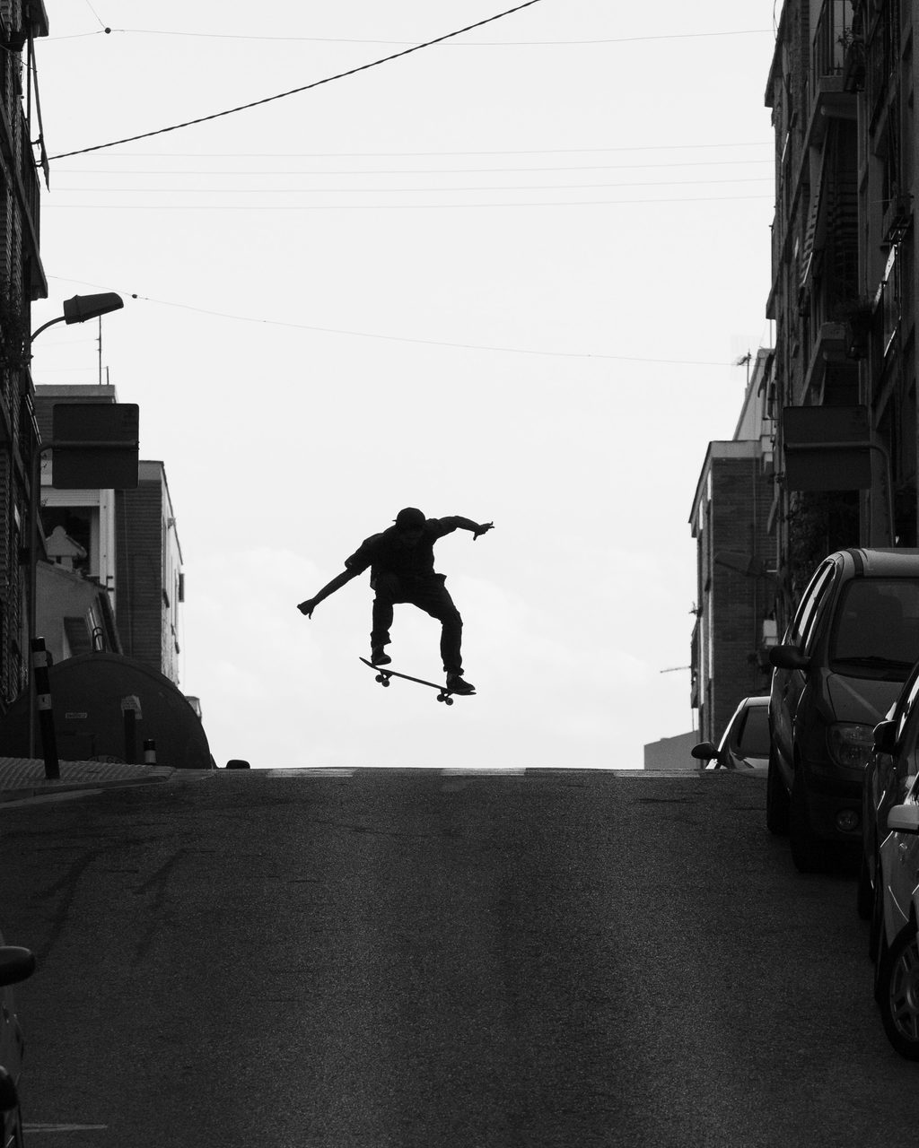 Silueta de un skater en pleno salto sobre una calle empinada de Burjassot, Valencia, con edificios y coches aparcados a ambos lados, en blanco y negro.