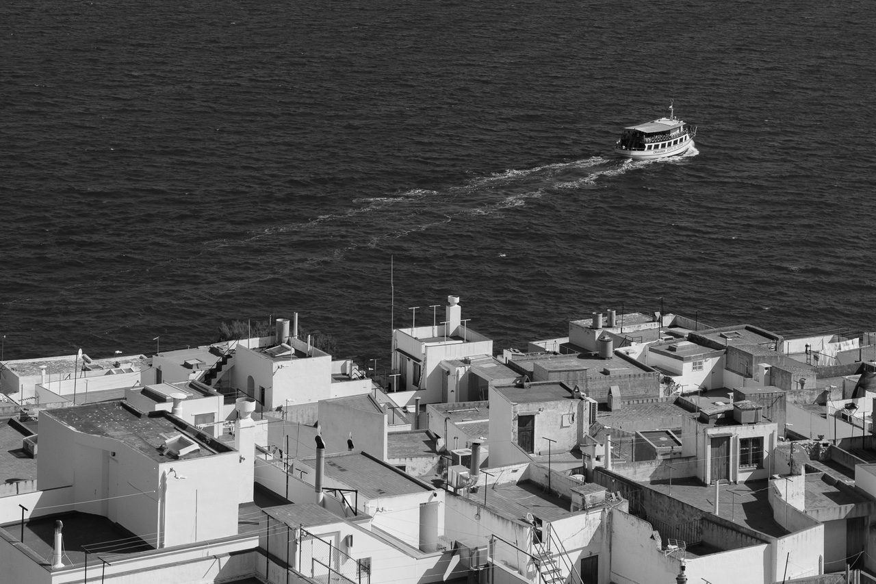 Tejados blancos del casco antiguo de Peñíscola vistos desde el castillo, con un barco navegando en el mar Mediterráneo al fondo, en blanco y negro.