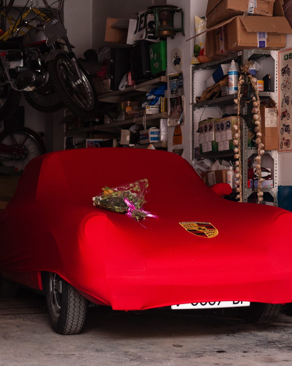 Porsche cubierto con una funda roja en un garaje de Madrid, con un ramo de flores sobre el capó y motos clásicas colgadas del techo entre herramientas.