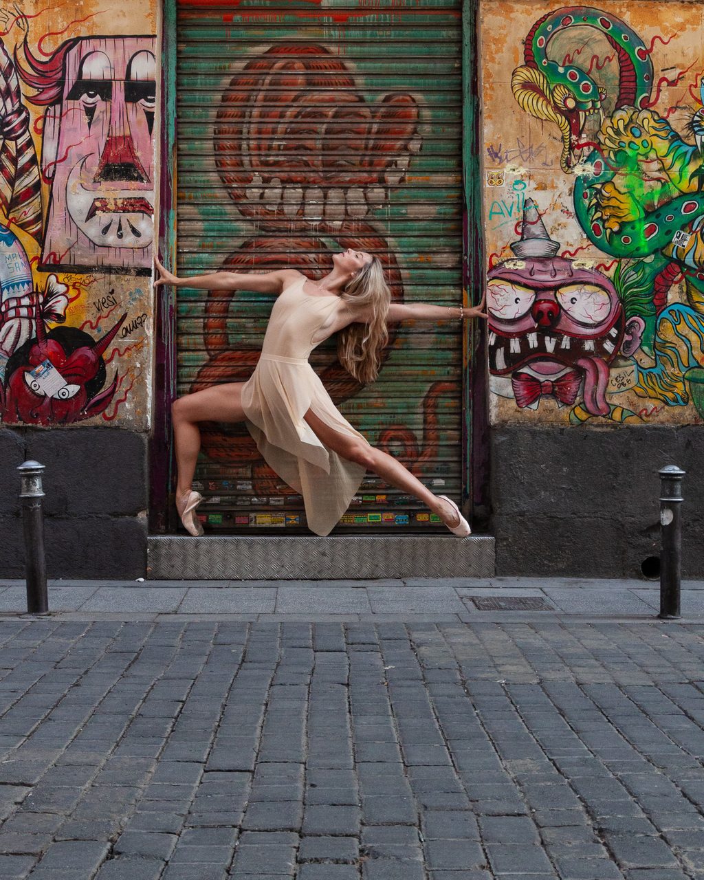 Cassandra Orefice en pose de punta con vestido claro frente a murales de street art y persianas pintadas en el barrio de Malasaña, Madrid.
