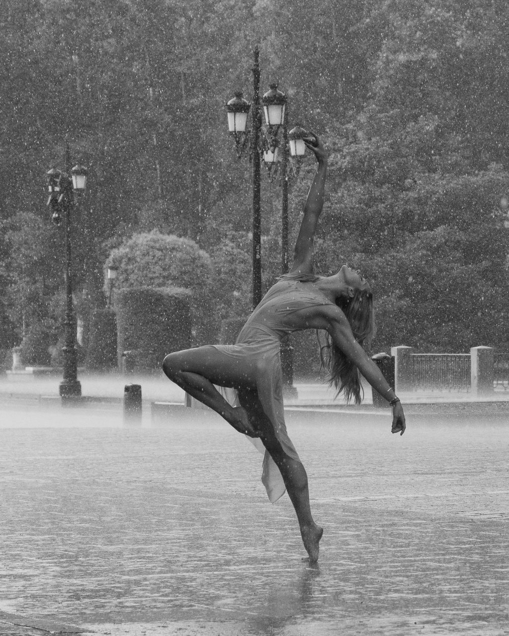 Cassandra Orefice bailando descalza bajo la lluvia junto al Palacio Real de Madrid en blanco y negro, con el cuerpo arqueado hacia atrás y una pierna elevada.
