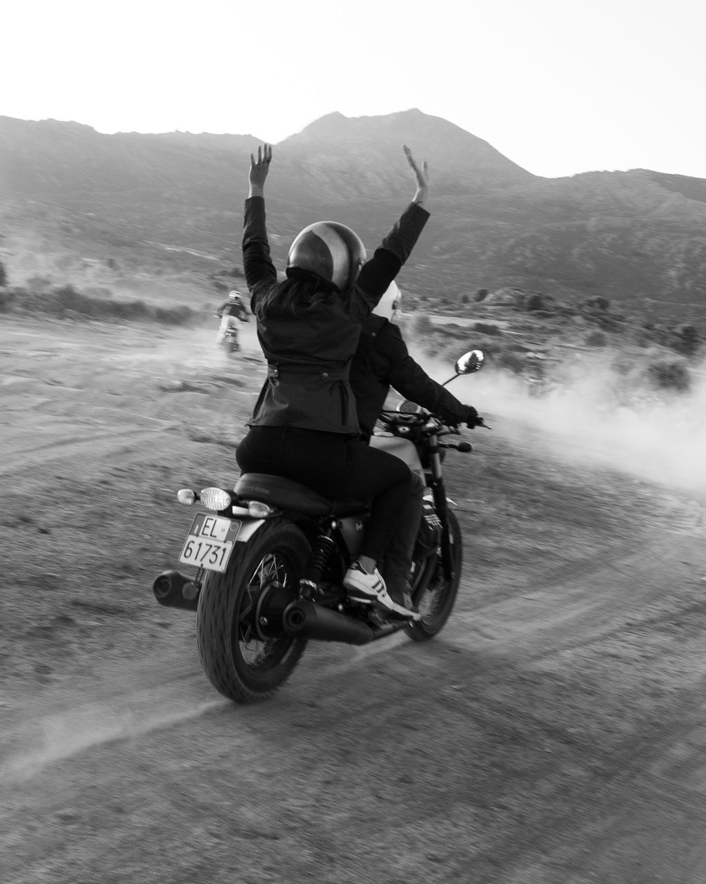 Moto Guzzi con dos personas recorriendo un camino de tierra en la Sierra de Madrid, con la pasajera levantando los brazos al cielo, en blanco y negro.