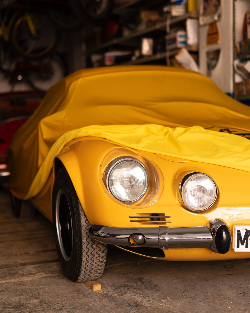 Frontal de un Alpine A110 parcialmente cubierto con una funda amarilla en un garaje de Madrid repleto de motos clásicas y herramientas.