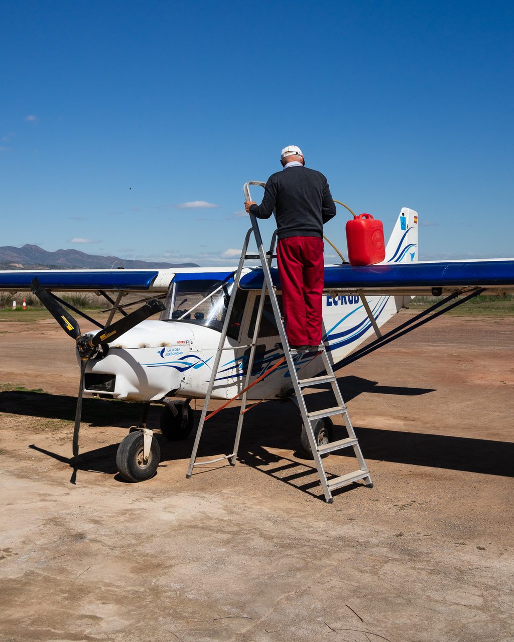 Piloto sobre una escalera de aluminio repostando un avión ultraligero con una garrafa roja en un aeródromo de Valencia, bajo un cielo azul despejado con montañas al fondo.

