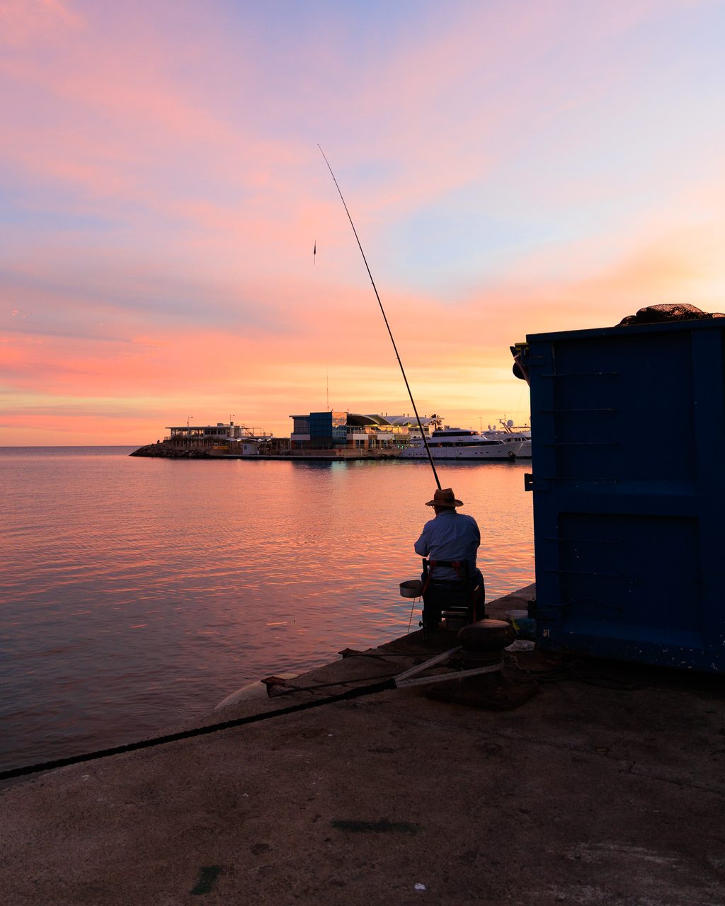 Hombre pescando al atardecer en el puerto de Burriana, Castellón, sentado en el borde del muelle con la caña elevada sobre un mar en calma de tonos rosados.
