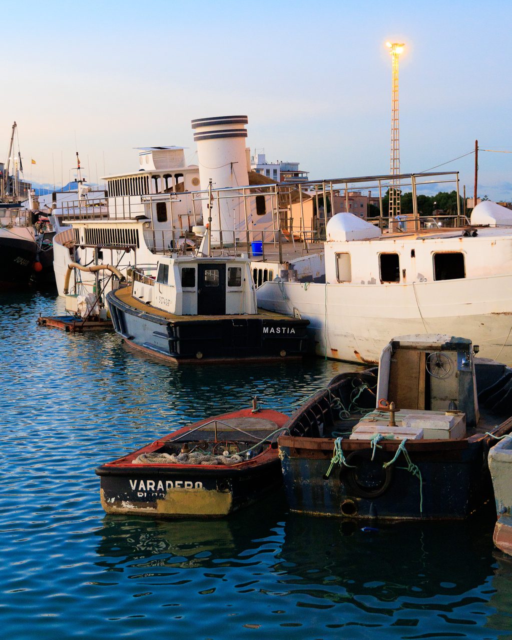 Embarcaciones de trabajo y remolcadores amarrados al anochecer en el puerto de Burriana, Castellón, con luz cálida de atardecer reflejada en el agua.