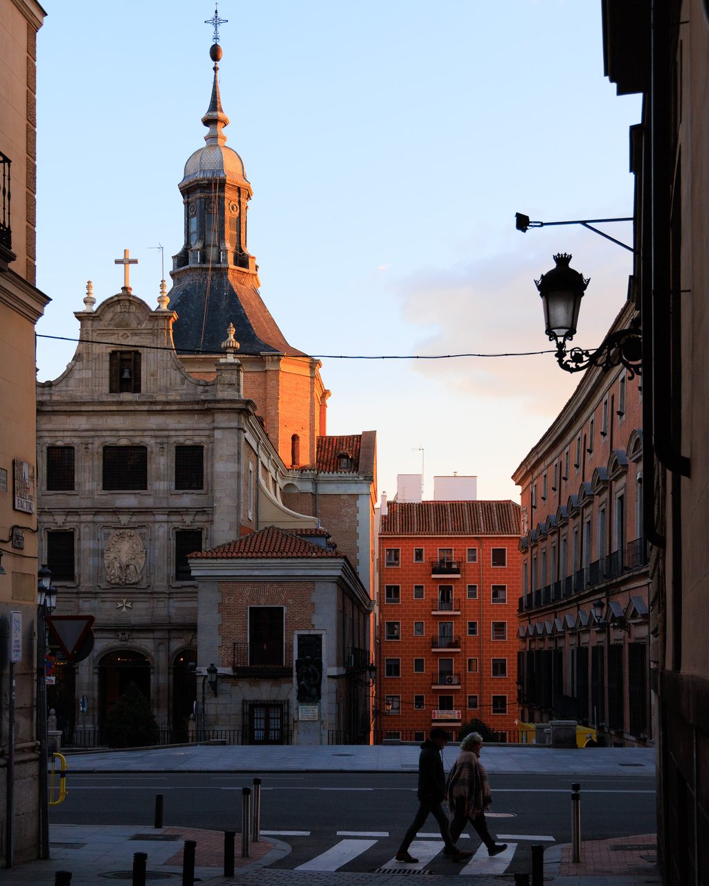 Dos siluetas cruzando un paso de peatones en Madrid al atardecer, con la luz dorada iluminando una iglesia barroca y fachadas históricas cerca del Palacio Real.