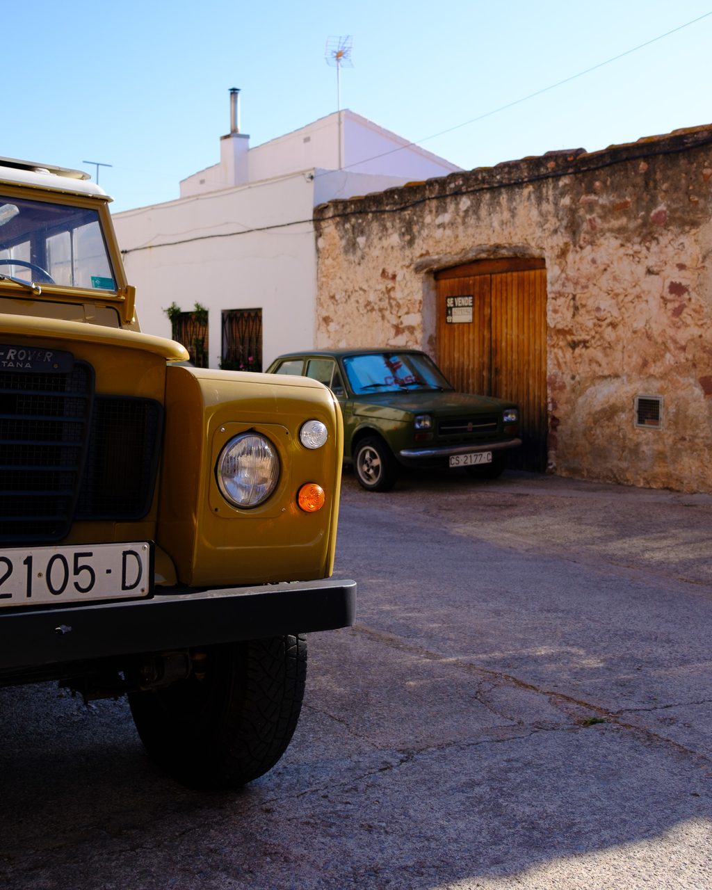 Land Rover Santana amarillo y un Seat 127 verde aparcados en una calle de piedra y fachadas encaladas en Vilafamés, Castellón, bajo un cielo despejado.