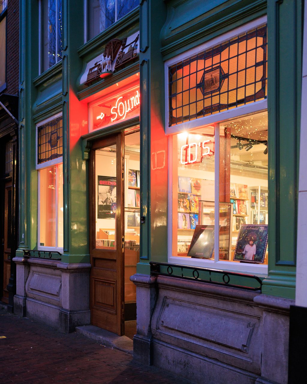La fachada de la tienda de música Sounds en Delft al anochecer, con el letrero de neón rojo encendido y un escaparate lleno de vinilos y carteles.