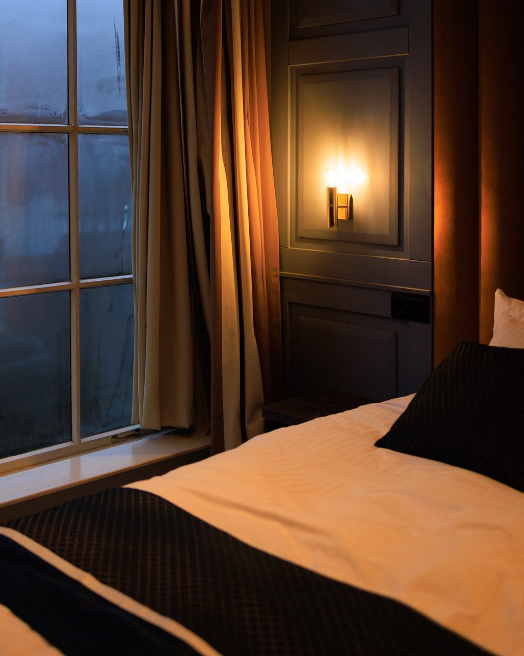 Interior de una habitación del hotel The Royal Bridges en Delft, con lámpara de pared encendida, cortinas elegantes y ventana empañada junto a la cama.