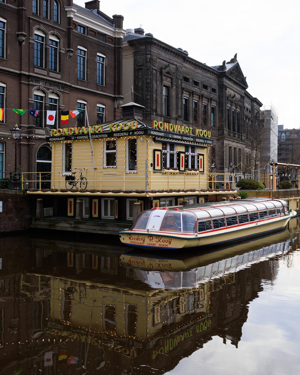 Barco de Rondvaart Kooij atracado en un canal de Ámsterdam, con fachadas históricas de ladrillo y luces del muelle reflejadas en el agua.