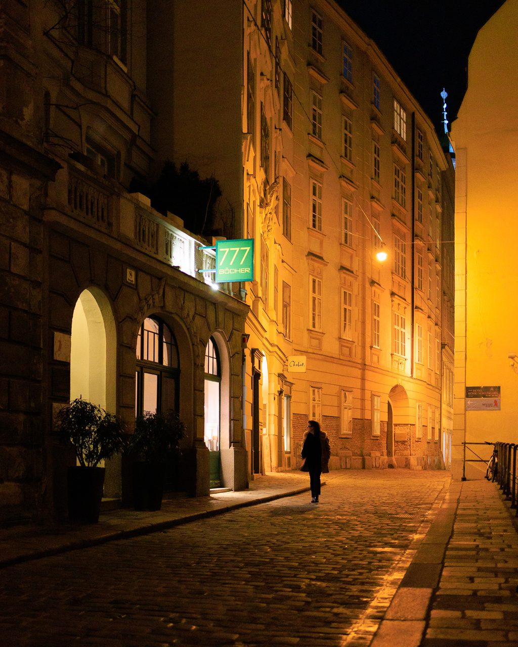 Una calle histórica de Viena iluminada por farolas doradas, con adoquines brillantes y una persona caminando sola entre edificios clásicos.