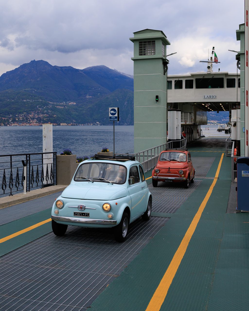 Dos Fiat 500 clásicos, uno azul y otro rojo, descendiendo por la rampa de un ferry en el Lago de Como, Bellagio, con montañas alpinas y cielo nublado al fondo.