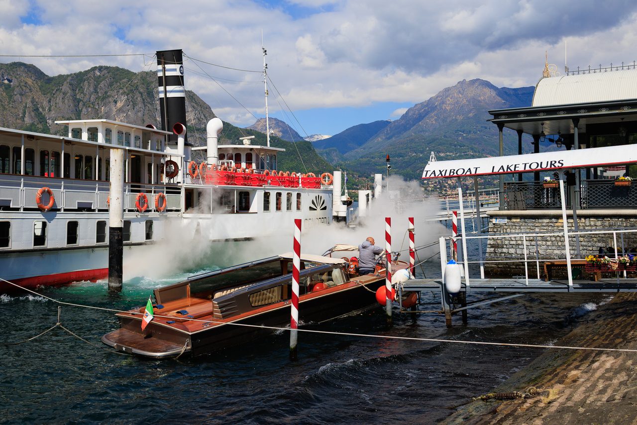 Muelle de Bellagio en el Lago de Como con un barco de vapor clásico emitiendo una nube de vapor blanco y un taxi boat de madera con bandera italiana, montañas alpinas al fondo.