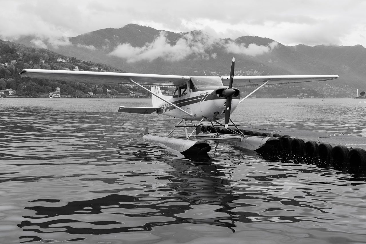 Un hidroavión del Aero Club Como sobre una plataforma flotante en el Lago de Como, con reflejos en el agua y montañas nubladas al fondo.