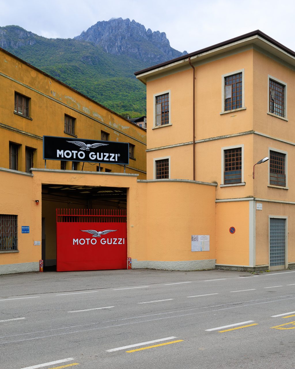 La entrada del Museo Moto Guzzi en Mandello del Lario, Lago de Como, con el portón rojo y logotipo alado enmarcado por edificios industriales y montañas del Prealpes lombardo.