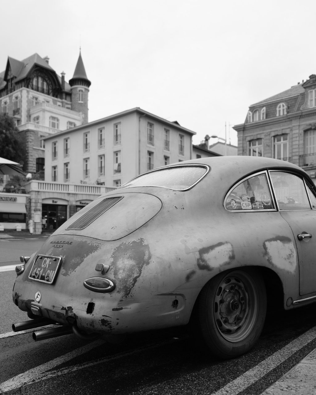 Un Porsche 356 envejecido aparcado en una calle de Biarritz, con edificios de arquitectura clásica y pavimento húmedo.