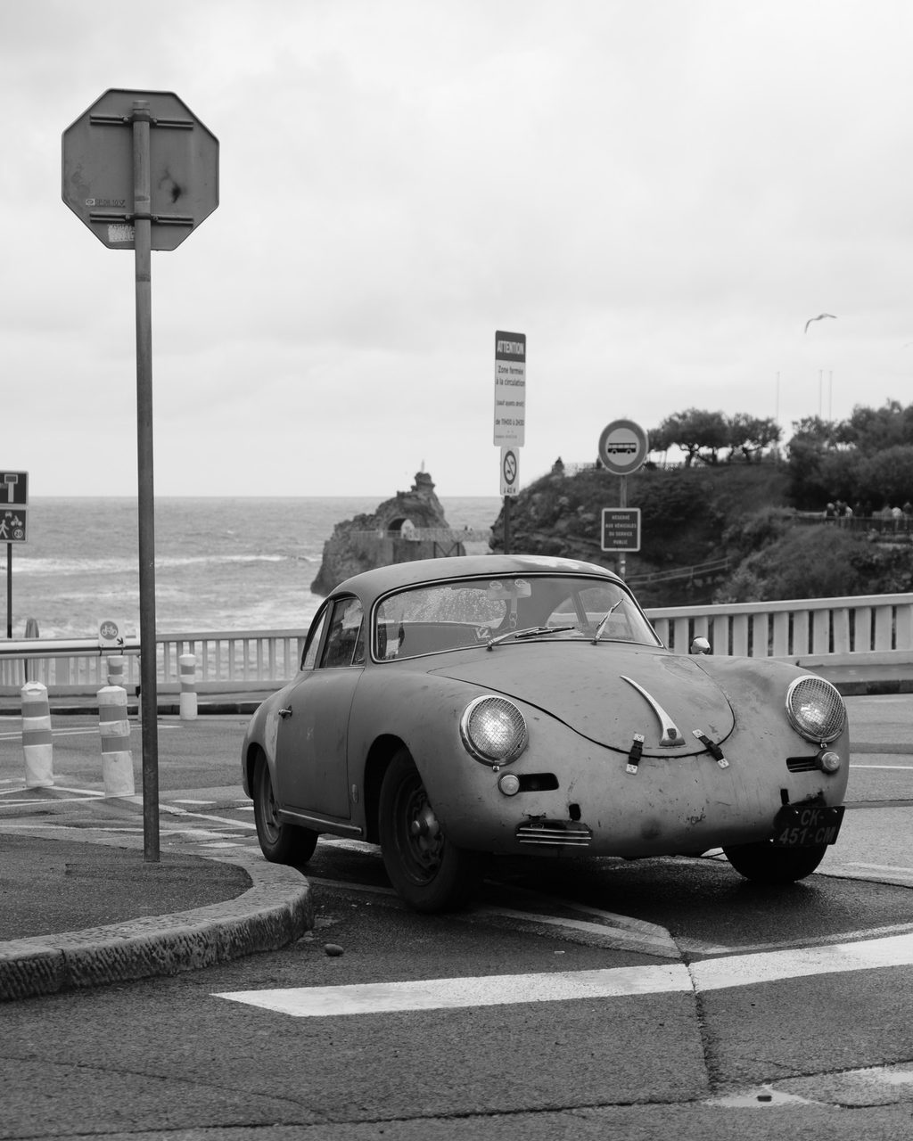 Un Porsche 356 envejecido aparcado junto a la costa atlántica de Biarritz, con acantilados, cielo nublado y una gaviota en vuelo.