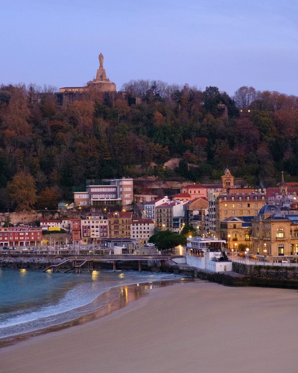 Amanecer en San Sebastián desde el Hotel de Londres y de Inglaterra, con la bahía de La Concha, la playa vacía y el monte Urgull coronado por el Sagrado Corazón bajo un cielo dorado.
