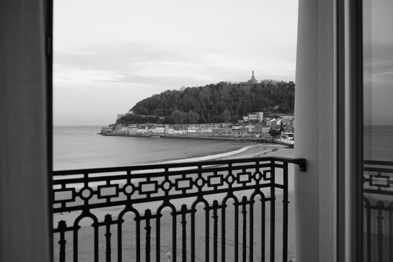 Amanecer en San Sebastián en blanco y negro enmarcado por el balcón de hierro forjado del Hotel de Londres y de Inglaterra, con la bahía de La Concha y el monte Urgull al fondo.
