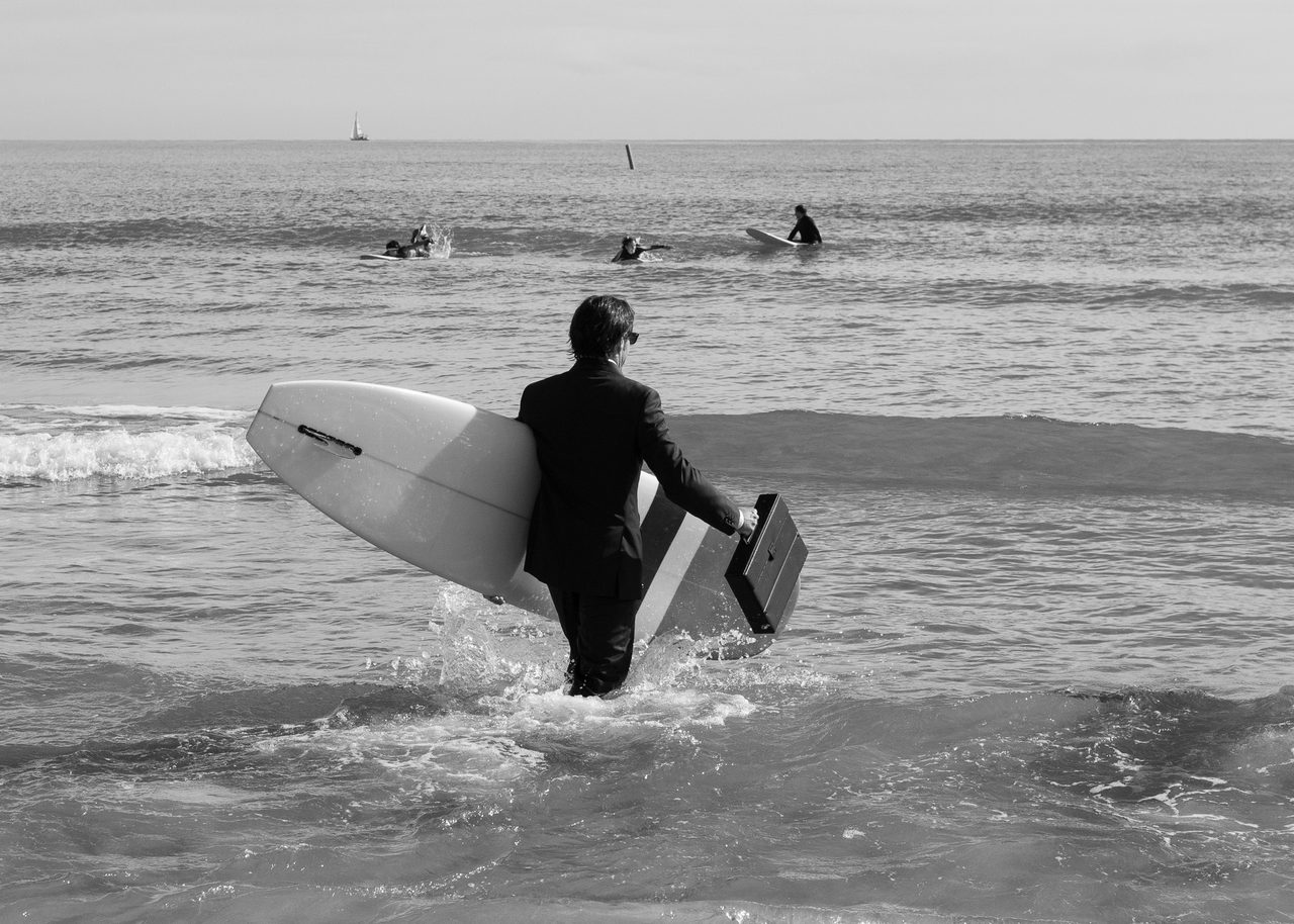 Persona vestida de ejecutivo con tabla de surf y maletín caminando hacia el mar durante el Festival Quasimoto en Burriana, Castellón, en blanco y negro.