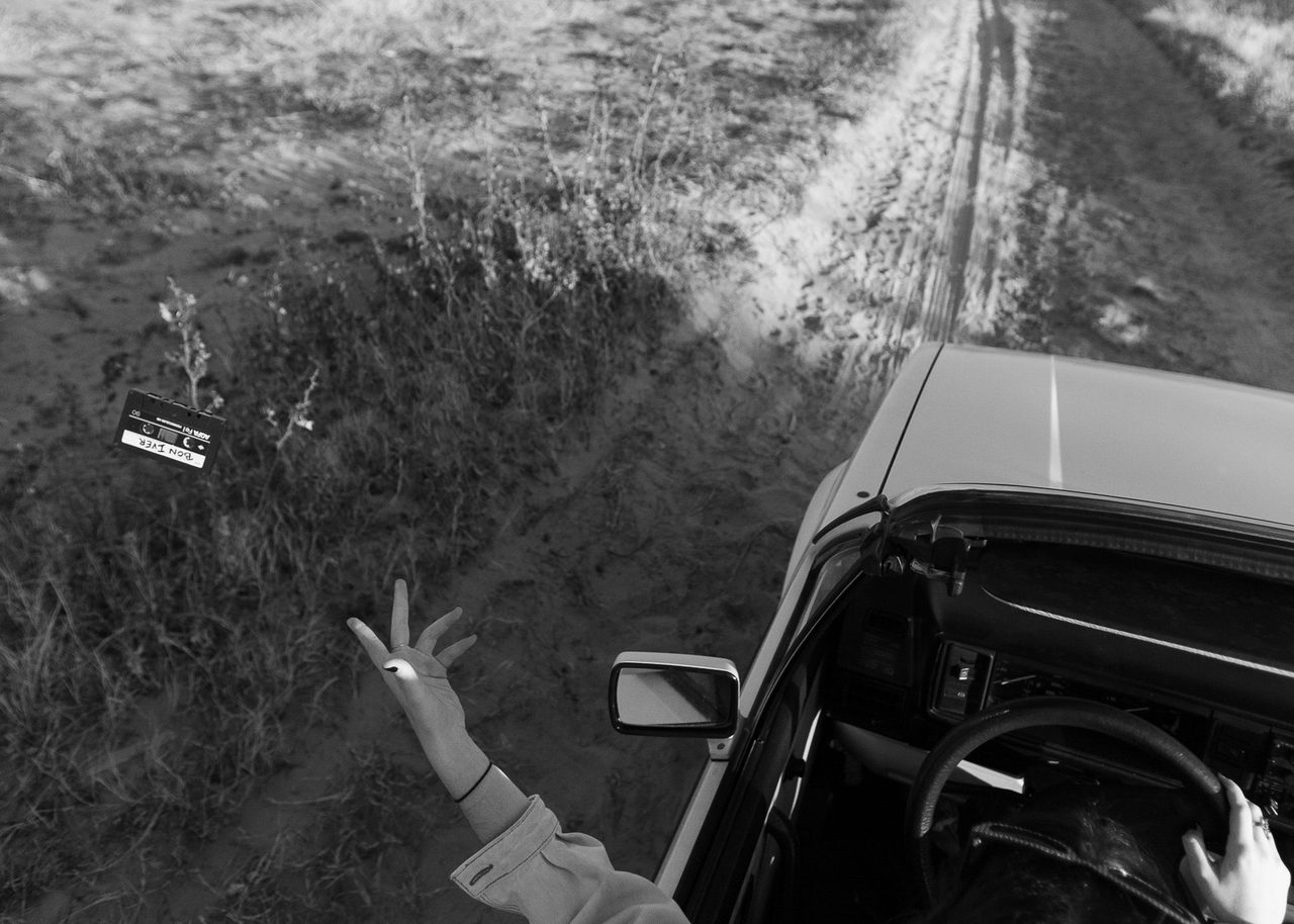 VW descapotable en un camino de arena junto a la playa de Burriana, con el conductor dejando caer una cinta de casete por la ventanilla, en blanco y negro.