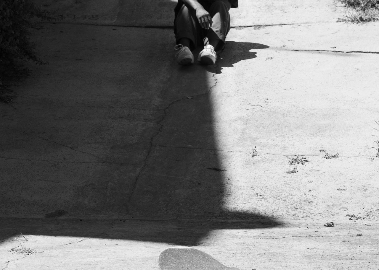 Skater sentado en la sombra de una rampa de hormigón en Castellón, con el monopatín iluminado por el sol más abajo en la pendiente, en blanco y negro.