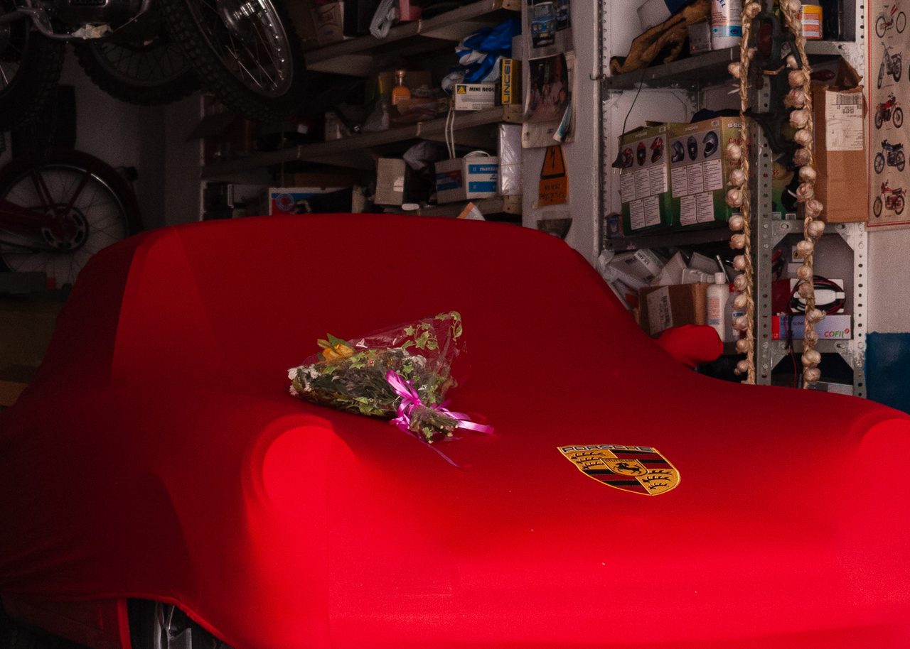 Porsche cubierto con una funda roja en un garaje de Madrid, con un ramo de flores sobre el capó y motos clásicas colgadas del techo entre herramientas.