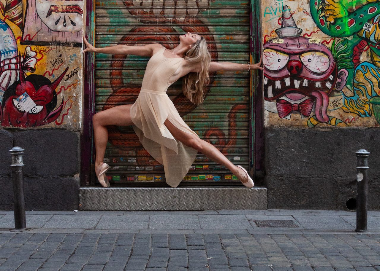 Cassandra Orefice en pose de punta con vestido claro frente a murales de street art y persianas pintadas en el barrio de Malasaña, Madrid.
