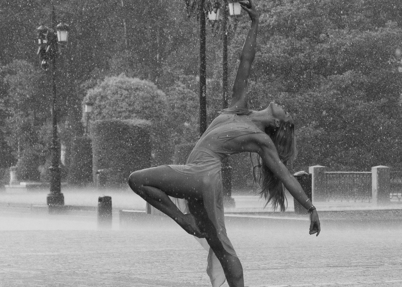 Cassandra Orefice bailando descalza bajo la lluvia junto al Palacio Real de Madrid en blanco y negro, con el cuerpo arqueado hacia atrás y una pierna elevada.
