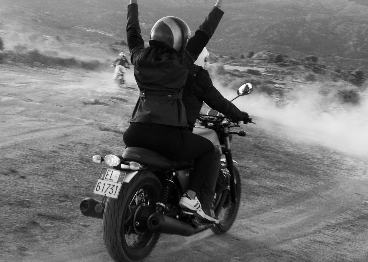 Moto Guzzi con dos personas recorriendo un camino de tierra en la Sierra de Madrid, con la pasajera levantando los brazos al cielo, en blanco y negro.