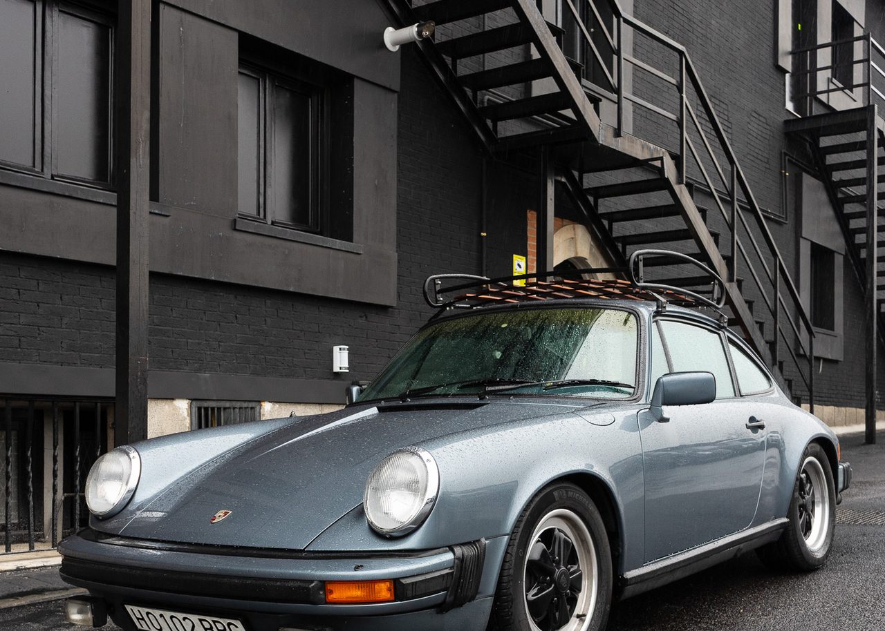 Porsche clásico con gotas de lluvia sobre la carrocería estacionado en una calle mojada de Madrid junto a una fachada industrial oscura.
