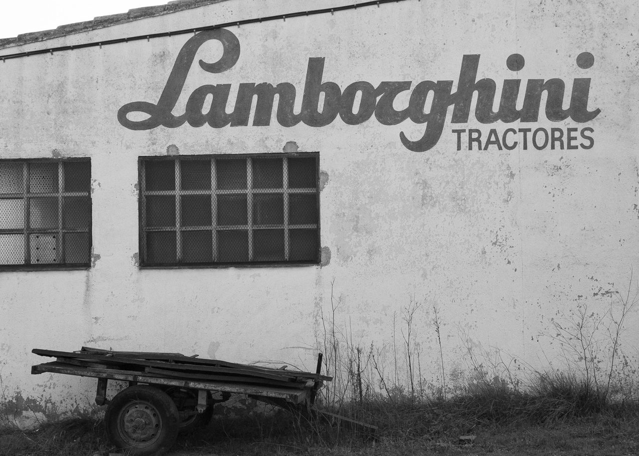 La fachada abandonada de un taller de Lamborghini Tractores en Castellón, con rótulo original visible y un remolque en desuso apoyado contra el muro agrietado.
