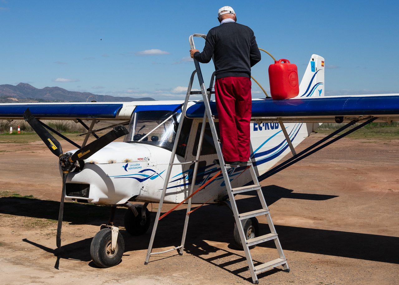 Piloto sobre una escalera de aluminio repostando un avión ultraligero con una garrafa roja en un aeródromo de Valencia, bajo un cielo azul despejado con montañas al fondo.