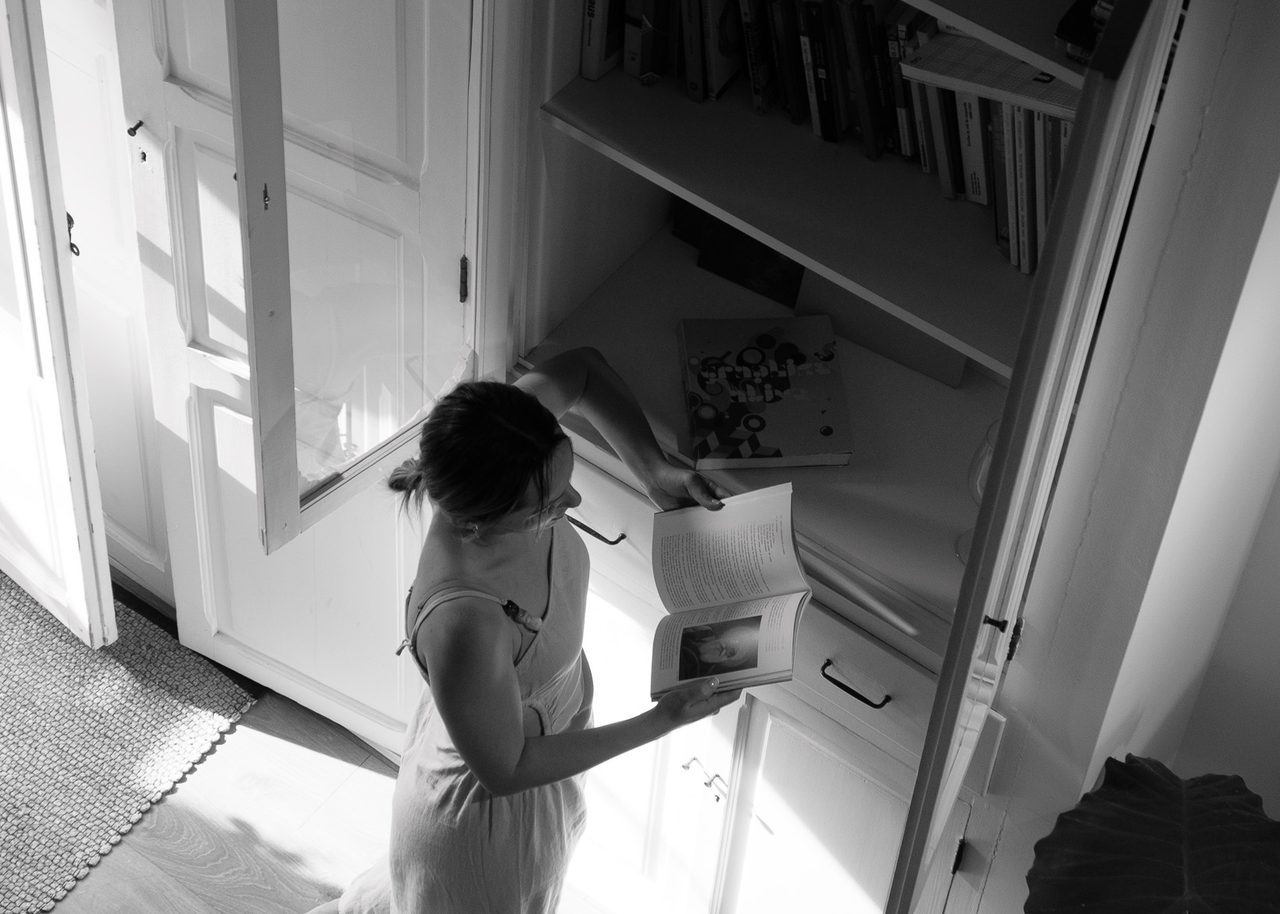 Una mujer descalza leyendo un libro en el interior de una antigua casa de pescadores del Cabanyal, Valencia, con luz natural entrando por las puertas de madera.