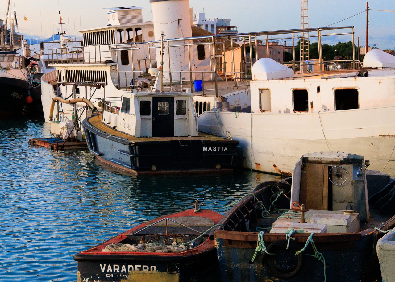 Embarcaciones de trabajo y remolcadores amarrados al anochecer en el puerto de Burriana, Castellón, con luz cálida de atardecer reflejada en el agua.
