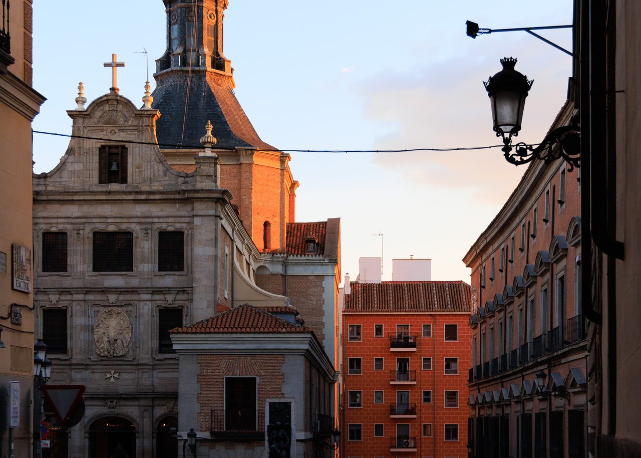 Dos siluetas cruzando un paso de peatones en Madrid al atardecer, con la luz dorada iluminando una iglesia barroca y fachadas históricas cerca del Palacio Real.