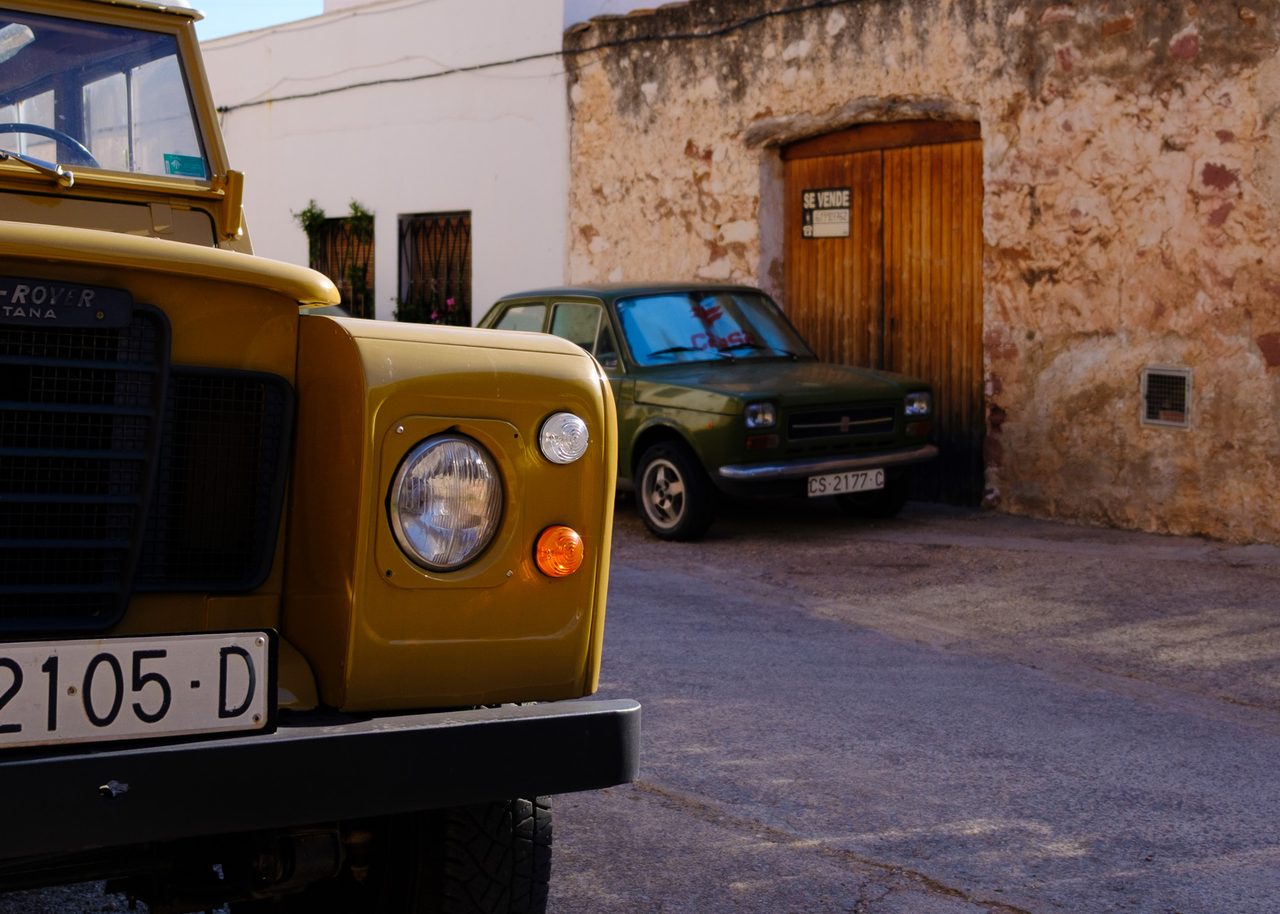 Land Rover Santana amarillo y un Seat 127 verde aparcados en una calle de piedra y fachadas encaladas en Vilafamés, Castellón, bajo un cielo despejado.

