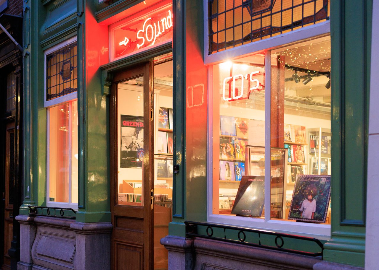 La fachada de la tienda de música Sounds en Delft al anochecer, con el letrero de neón rojo encendido y un escaparate lleno de vinilos y carteles.

