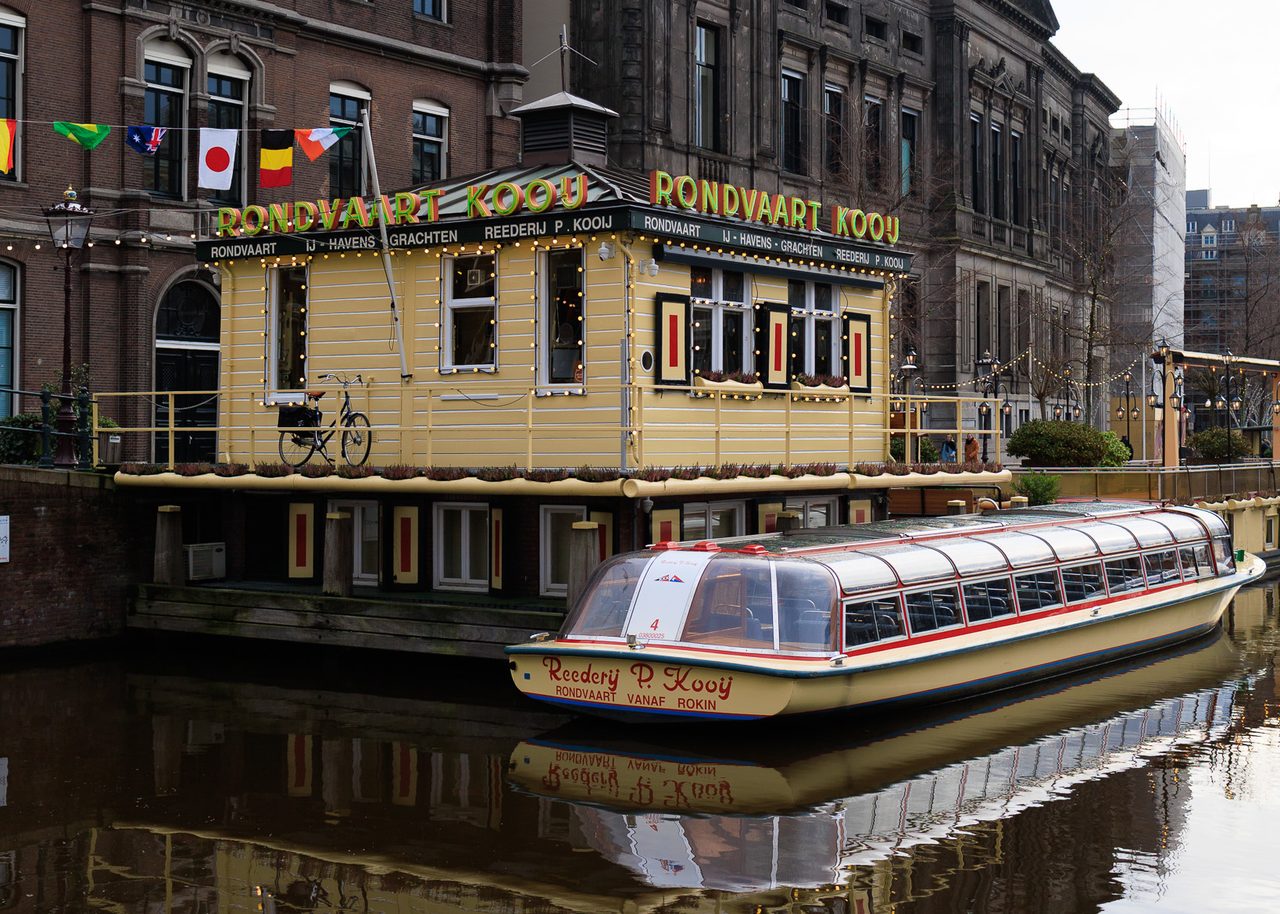 Barco de Rondvaart Kooij atracado en un canal de Ámsterdam, con fachadas históricas de ladrillo y luces del muelle reflejadas en el agua.
