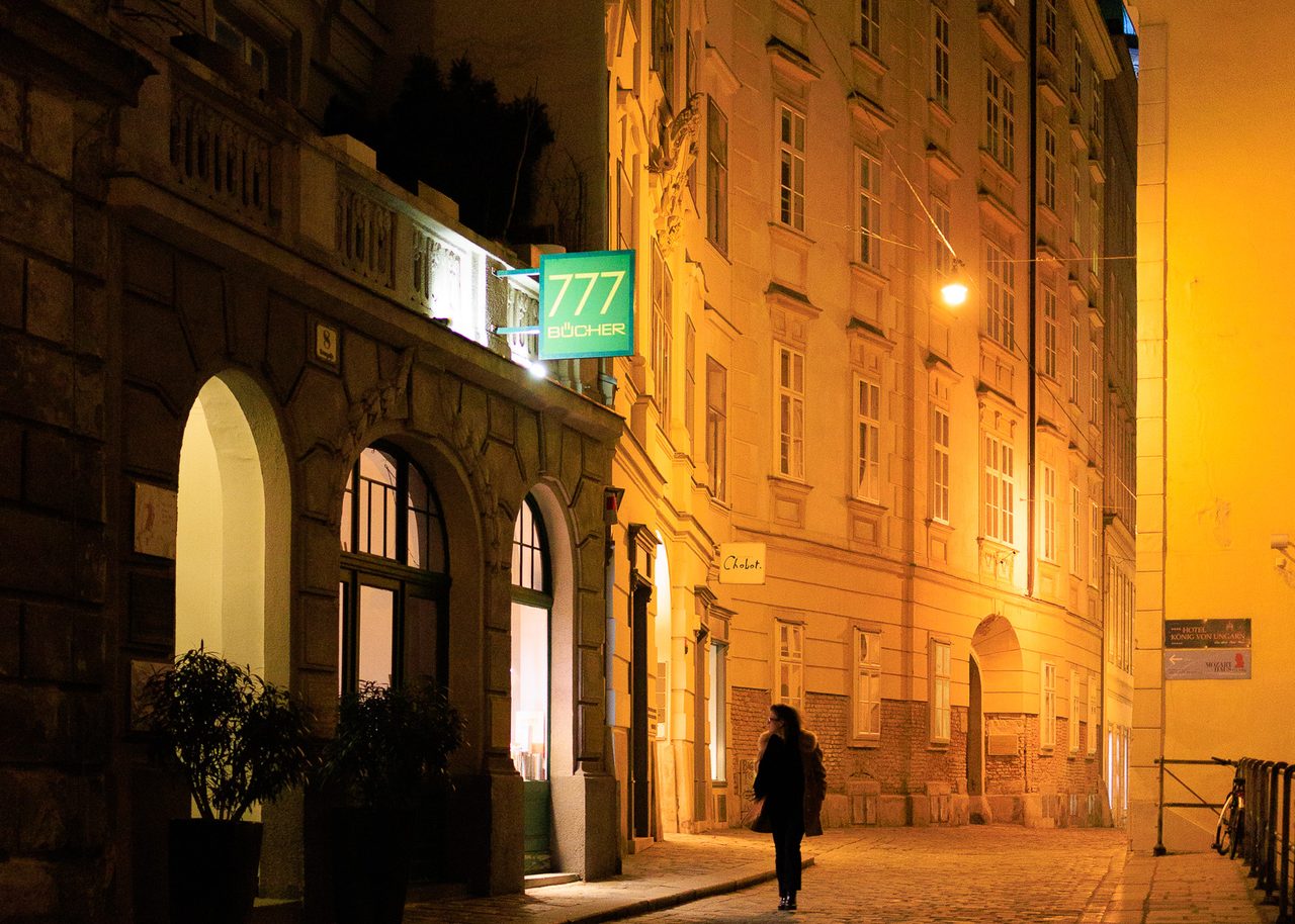 Una calle histórica de Viena iluminada por farolas doradas, con adoquines brillantes y una persona caminando sola entre edificios clásicos.