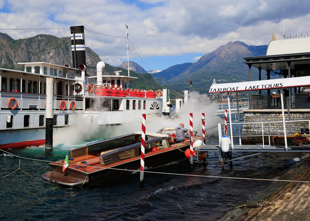 Muelle de Bellagio en el Lago de Como con un barco de vapor clásico emitiendo una nube de vapor blanco y un taxi boat de madera con bandera italiana, montañas alpinas al fondo.
