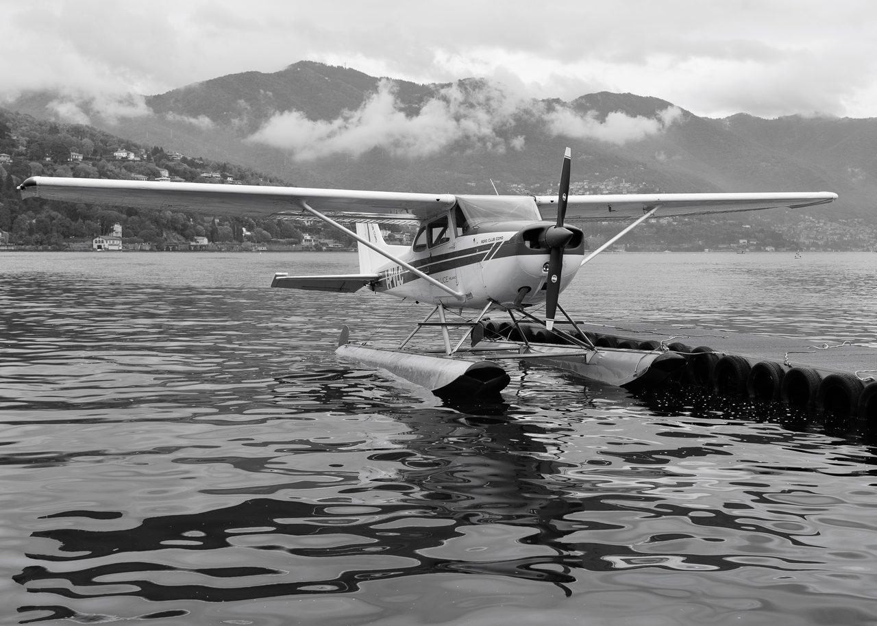 Un hidroavión del Aero Club Como sobre una plataforma flotante en el Lago de Como, con reflejos en el agua y montañas nubladas al fondo.