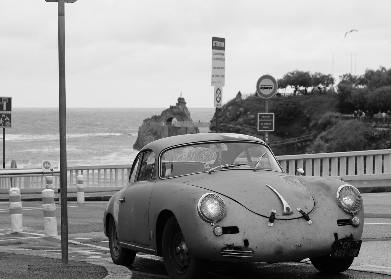 Un Porsche 356 envejecido aparcado junto a la costa atlántica de Biarritz, con acantilados, cielo nublado y una gaviota en vuelo.
