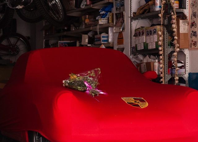 Porsche cubierto con funda roja y flores sobre el capó en un garaje con motos clásicas