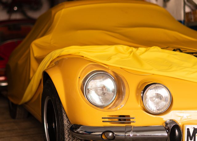 Frontal de un Alpine A110 asomando bajo una funda amarilla en un garaje repleto de motos